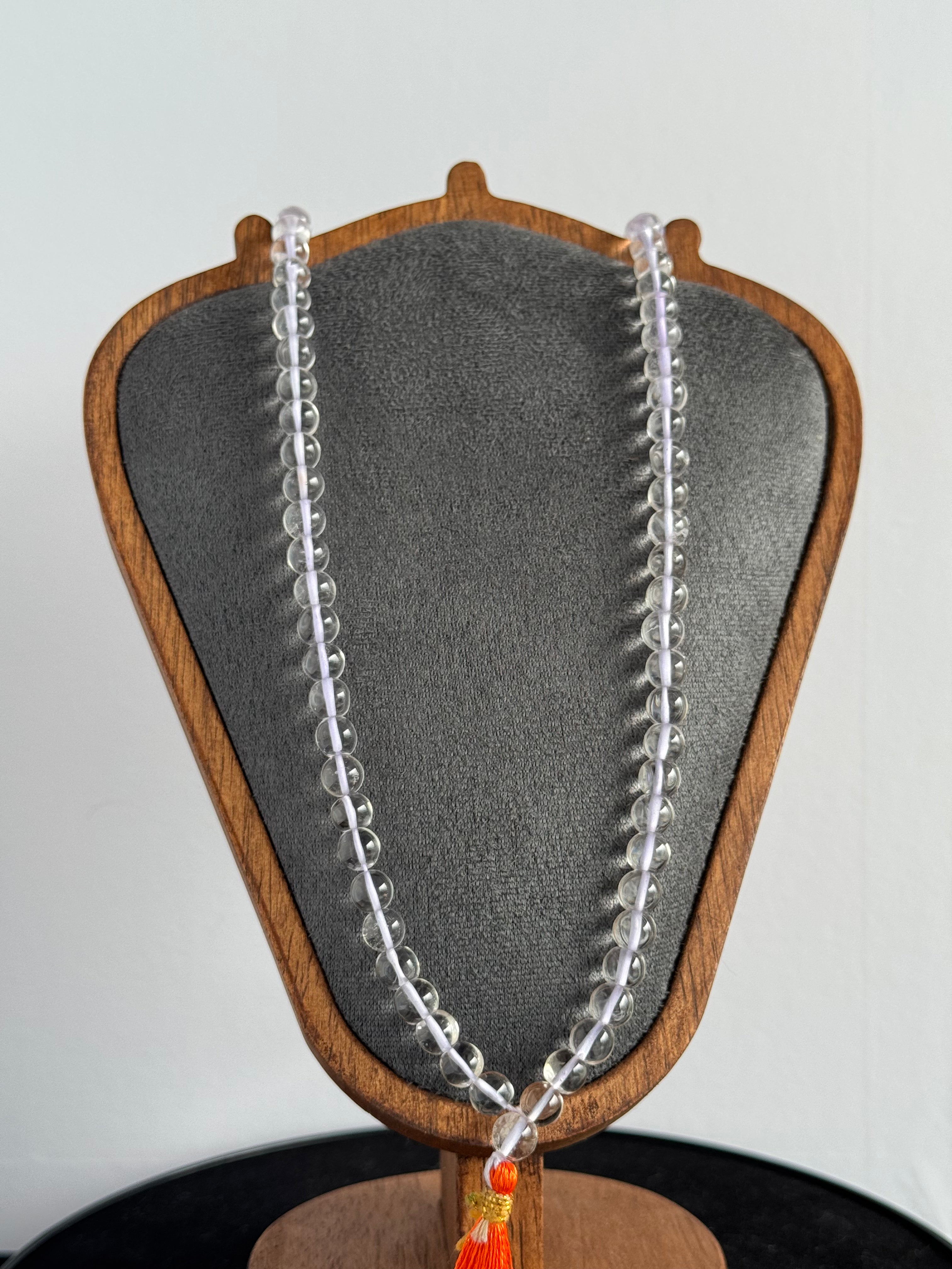 Real and pure sphatik mala/necklace. Made with authentic Himalayan quartz/Sphatik exclusively for Damru India( Damru.Shop)  A clear beaded necklace draped over a gray velvet jewelry bust with a wooden frame, shown against a plain light background.